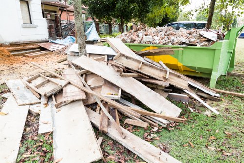 A pile of discarded wooden window frames, trims, and flat panels with visible grain and painted surfaces, positioned on a grassy area near a green skip filled with construction debris, including bricks and rubble, on the driveway of a residential property. The window frames and panels are weathered, with some showing chipped paint and rough edges, indicating prior use. The scene includes a background of trees, a white building, and other construction materials, suggesting an ongoing house clearance or renovation project in a suburban or residential area, possibly in Guildford. The image reflects typical rubbish removal tasks handled by House Clearance Guildford, featuring waste materials associated with building refurbishment or demolition activities, with natural daylight illuminating the scene and casting soft shadows on the grass and rubbish.