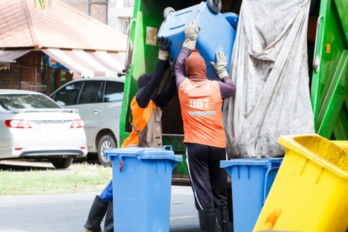 Two waste management workers dressed in high-visibility orange vests and black rubber boots are lifting a large blue plastic bin into a green rubbish collection vehicle parked on a residential street. One worker is wearing blue gloves, while the other has grey gloves, both focused on carefully loading the bin. The scene shows a typical urban environment with a parked silver car and a building with a shopfront and striped awning in the background. To the right, a yellow recycling bin is visible, positioned on the pavement alongside two similar blue bins. The waste collection process involves careful handling of the bin, supported by the vehicle's open rear side, which is partially covered with a grey tarp. The setting appears to be during daytime, with natural light illuminating the scene, emphasizing the outdoor context of the rubbish removal service in Guildford, which aligns with local waste management activities. The overall scene reflects professional rubbish collection, emphasizing the importance of proper waste handling on a typical residential street.