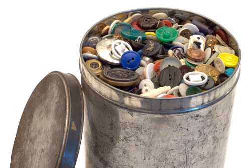 A round metal tin container filled with an assortment of mixed buttons in various sizes, shapes, and colours, including black, white, blue, green, and shades of brown, with some buttons made of plastic and others of metal. The container, which has a slightly weathered appearance and a partially visible removable lid placed beside it, is situated on a plain white background. The buttons are tightly packed and spilled over slightly at the top edge, demonstrating an organised collection or stored selection of buttons. This scene is relevant to rubbish removal services provided by House Clearance Guildford, especially when clearing out sewing or craft supplies from properties in the Guildford area, within the GU postcode district, or nearby towns in Surrey.