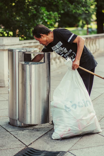 A street scene during daytime featuring a red waste collection vehicle parked on the side of the road with its rear compactor open, revealing the emptying mechanism. A worker wearing a red vest with y