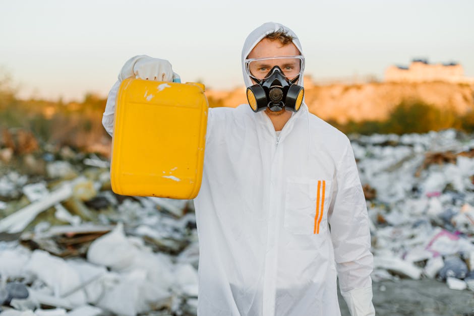 A light blue disposable face mask made of non-woven fabric lies discarded among dry, brown plant debris on the ground, with a partial view of a red gardening glove visible nearby. The scene appears to